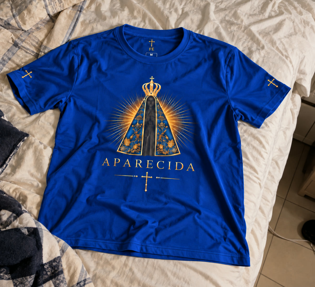 Camiseta de Nossa Senhora Aparecida aberta na cama, com a estampa da frente e as cruzes douradas nas mangas.